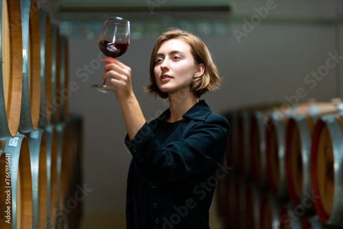 Quadro su tela Professional Caucasian woman sommelier tasting and sniffing red wine in wine glass at wine cellar with wooden barrel