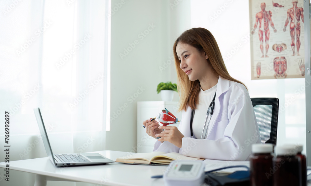 Young Asian doctor in white medical uniform with stethoscope using laptop computer video chatting with patient at table in health clinic or hospital