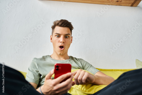 surprised student in his 20s at home sitting on yellow couch and scrolling in social media