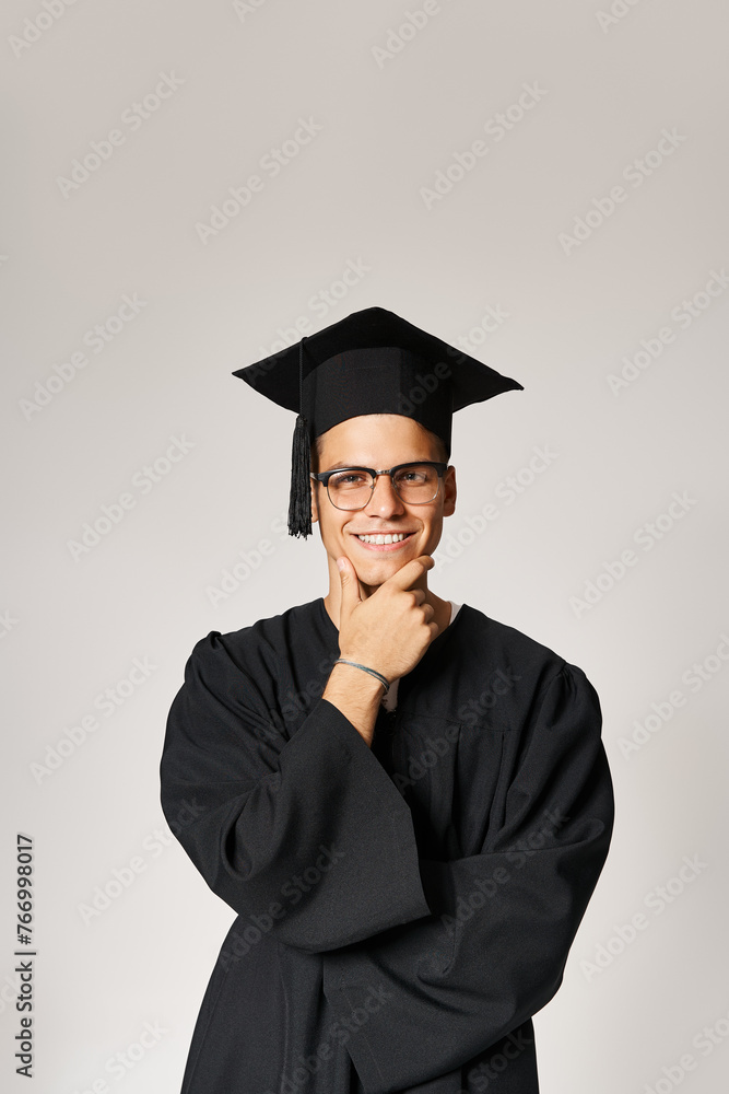 smiling student in graduate outfit and vision glasses touching hand to jawline on grey background smiling student in graduate outfit and vision glasses touching hand to jawline on grey background