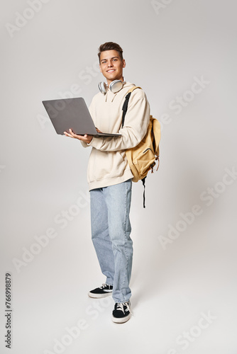 young student in headphones standing with backpack and networking to laptop against grey background