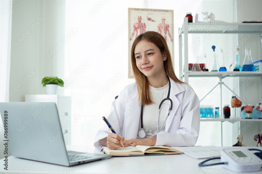 Young Asian doctor in white medical uniform with stethoscope using laptop computer video chatting with patient at table in health clinic or hospital Young Asian doctor in white medical uniform with stethoscope using laptop computer video chatting with patient at table in health clinic or hospital