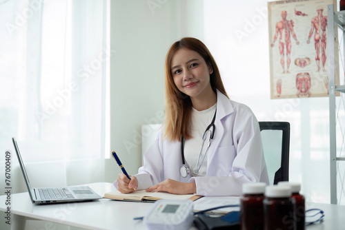 Young Asian woman or female medical staff sitting smiling, arms looking happily at camera, working on table in clinic office in telehealth, telemedicine, consulting online service.