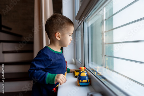 Little boy stands near the window and looks out the open window with toy car