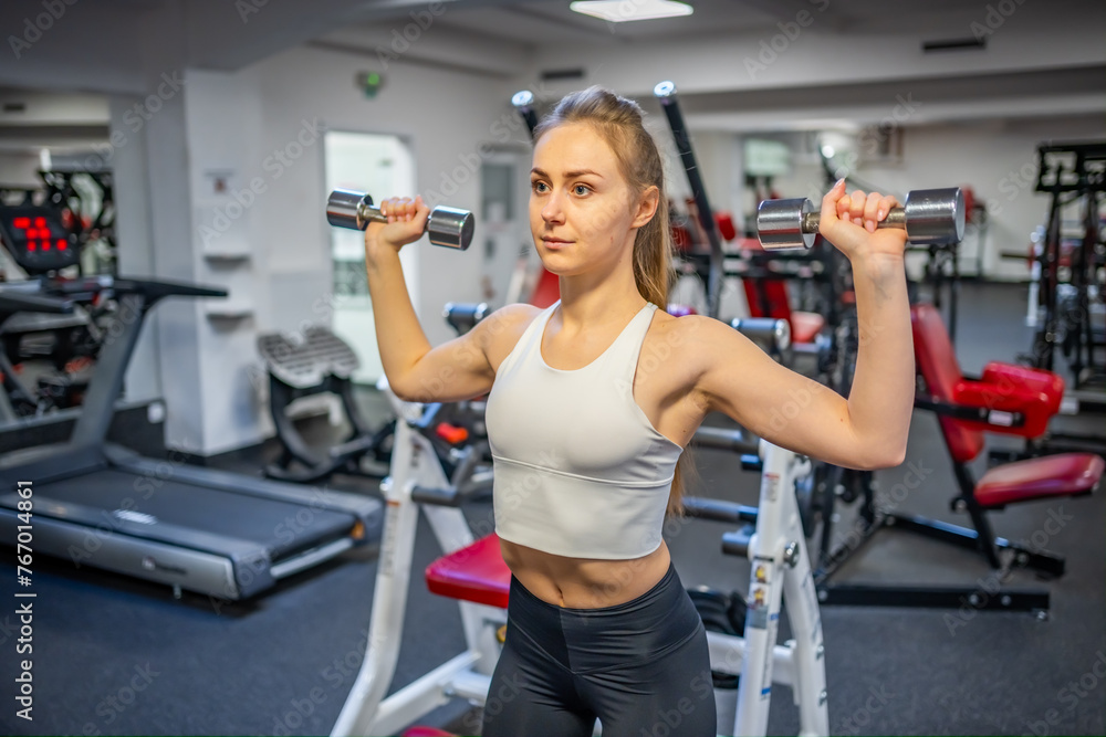 Young woman holds dumbbells in her hands and works out in the gym performing an exercise - obrazy, fototapety, plakaty 