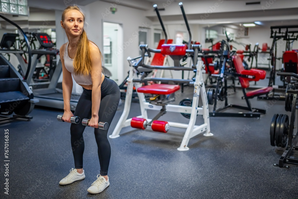 Young woman holds dumbbells in her hands and works out in the gym performing an exercise Young woman holds dumbbells in her hands and works out in the gym performing an exercise