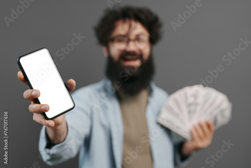 An exuberant bearded man holds out a smartphone with a blank screen and a fan of cash, expressing success and digital finance opportunities against a neutral gray backdrop.