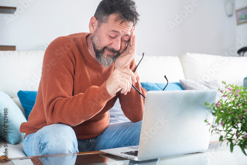 Tired sleepy mature freelancer man sitting on sofa at work laptop and touching glasses. Overwork and overtime small business online. People using too much computer laptop. Home office business photo
