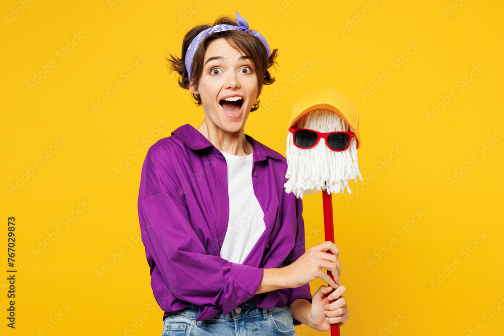 Young shocked excited woman wear purple shirt casual clothes do housework tidy up hold in hand mop in hat and glasses kidding isolated on plain yellow background studio portrait. Housekeeping concept. Young shocked excited woman wear purple shirt casual clothes do housework tidy up hold in hand mop in hat and glasses kidding isolated on plain yellow background studio portrait. Housekeeping concept.