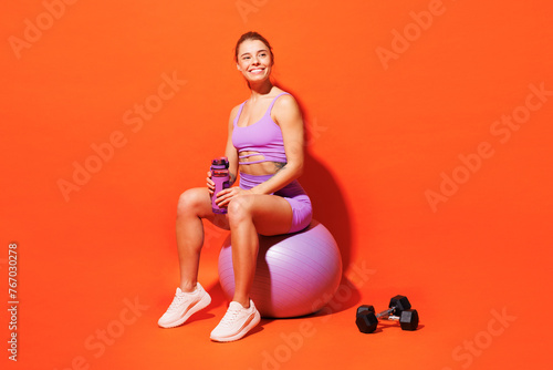 Full body young fitness trainer woman sportsman wears top shorts purple clothes in home gym sit on fit ball drink water look aside isolated on plain orange background. Workout sport fit abs concept.