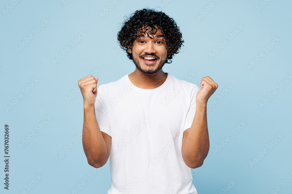 Young fun happy Indian man he wear white t-shirt casual clothes doing winner gesture celebrate clenching fists isolated on plain pastel light blue cyan background studio portrait. Lifestyle concept. - obrazy, fototapety, plakaty 