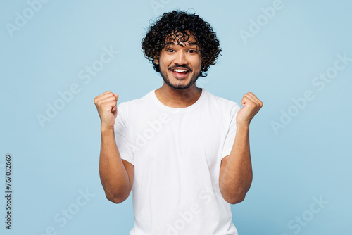 Young fun happy Indian man he wear white t-shirt casual clothes doing winner gesture celebrate clenching fists isolated on plain pastel light blue cyan background studio portrait. Lifestyle concept. © ViDi Studio
