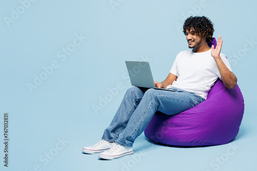 Full body young happy Indian man wear white t-shirt casual clothes sit in bag chair hold use work on laptop pc computer waving hand isolated on plain pastel light blue background. Lifestyle concept