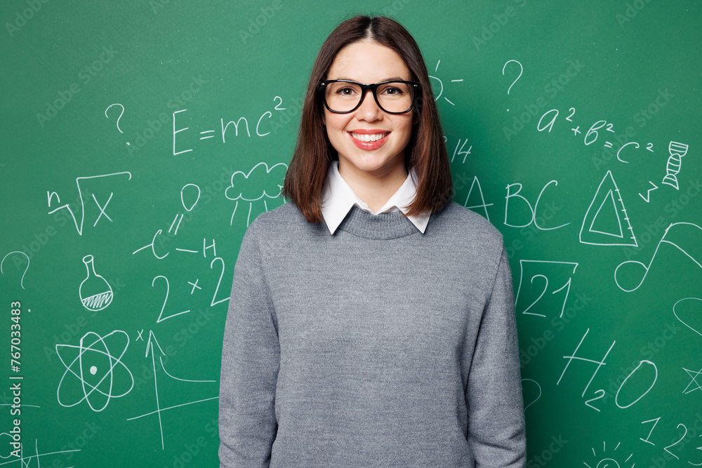 Young smiling happy cheerful smart teacher woman she wear grey casual shirt glasses looking camera isolated on green wall chalk blackboard background studio. Education in high school college concept.