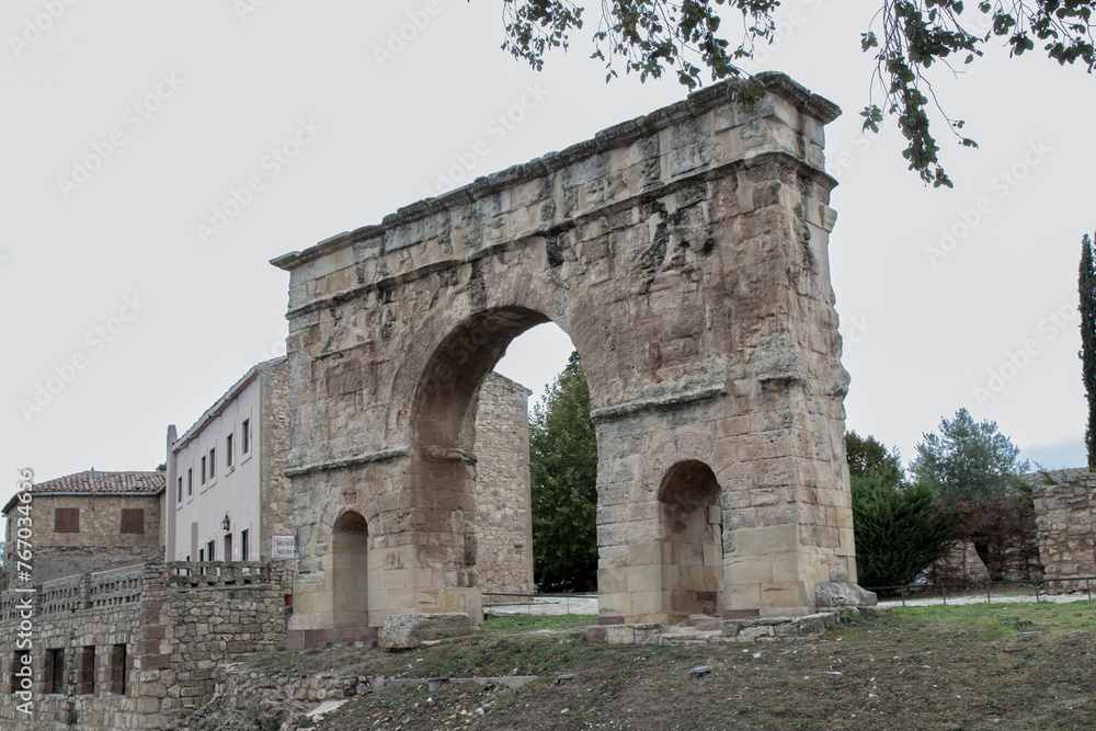 Ancient stone arch with two large openings, surrounded by trees and old buildings, under a cloudy sky