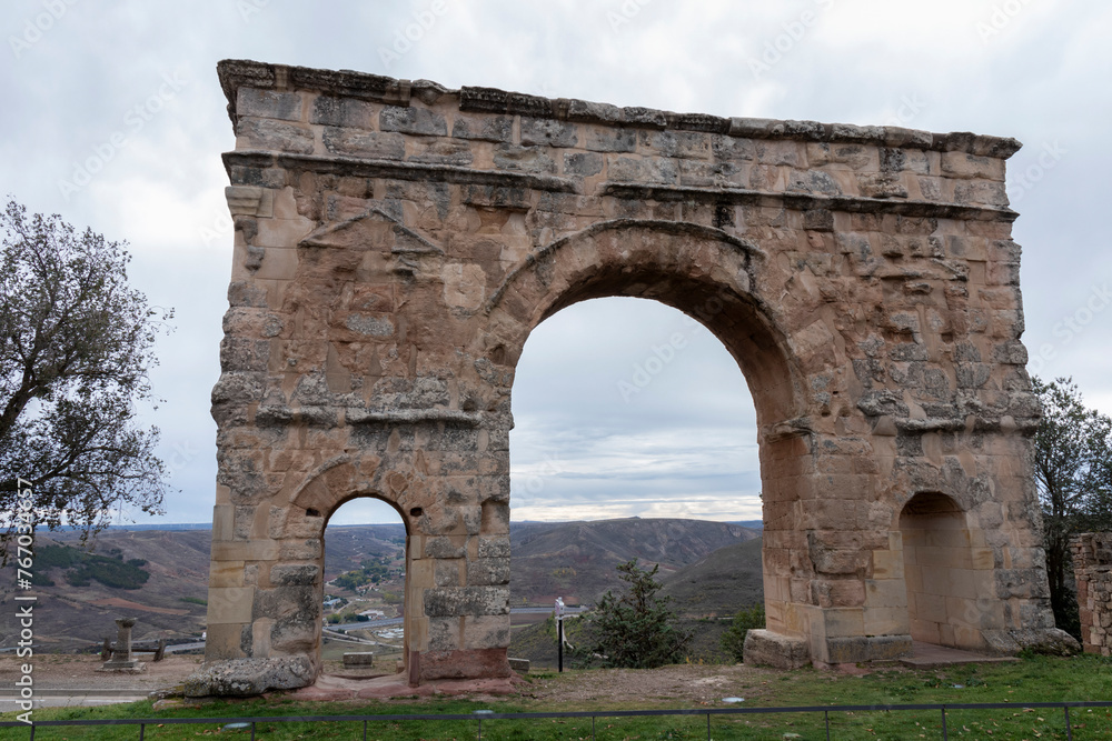 An ancient, weathered stone arch with two openings stands against a backdrop of a cloudy sky and distant landscape
