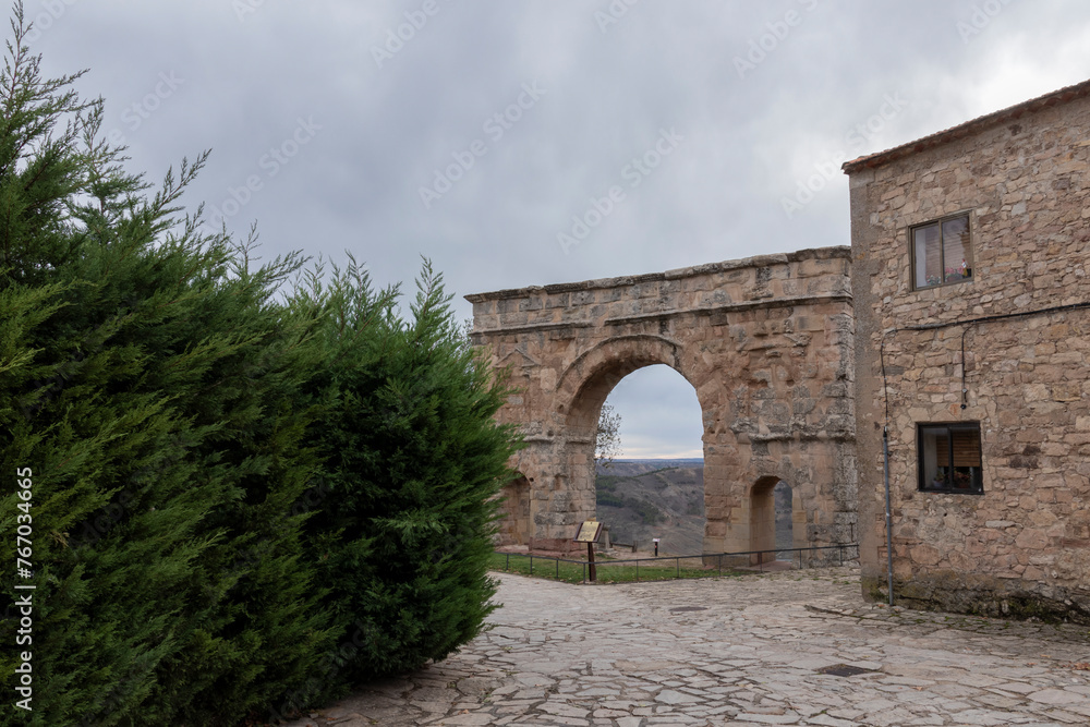 An old stone building with a large archway, adjacent to a green bush, under a cloudy sky