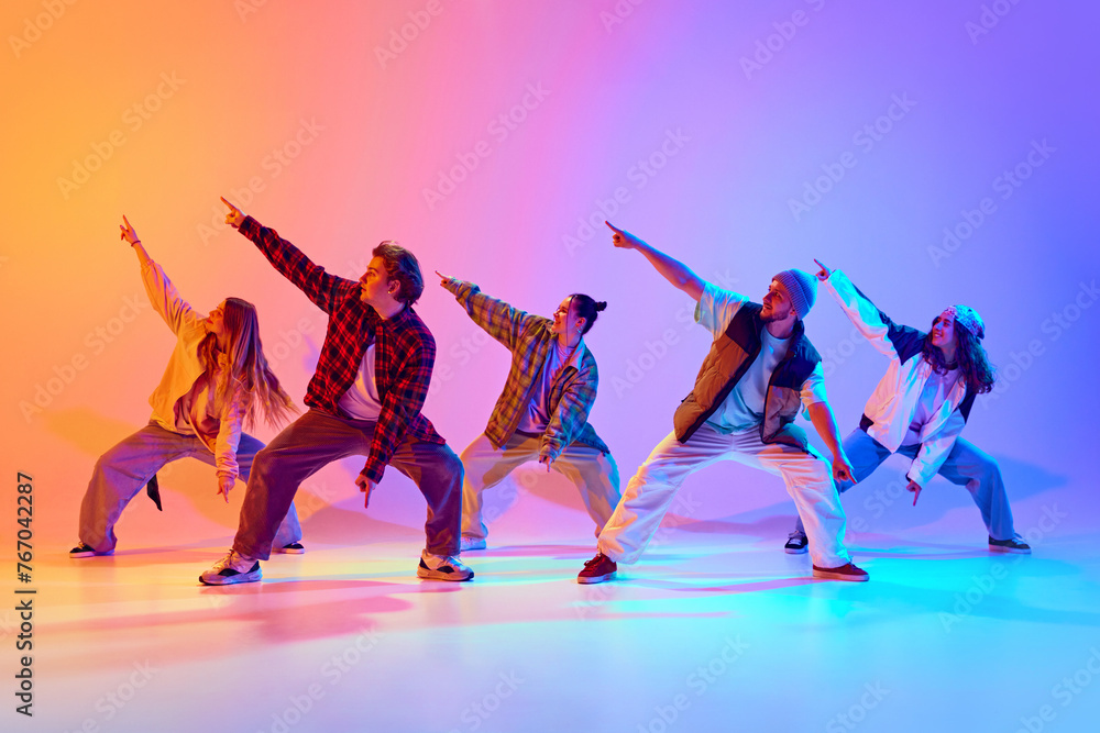 Fototapeta premium Group of five dancers in casual clothes performing with synchronized poses against gradient studio background in neon light. Concept of modern dance style, hobby, active lifestyle, youth culture