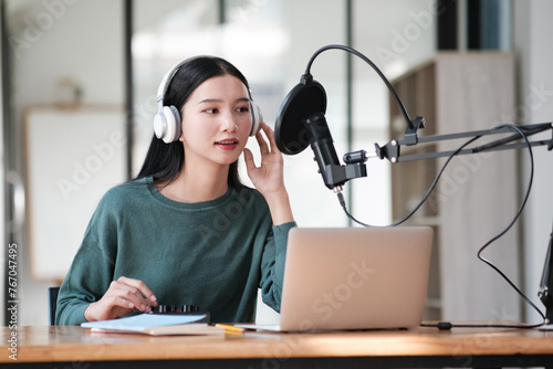 A woman wearing headphones is sitting at a desk with a laptop and a microphone. She is recording a podcast or a voiceover