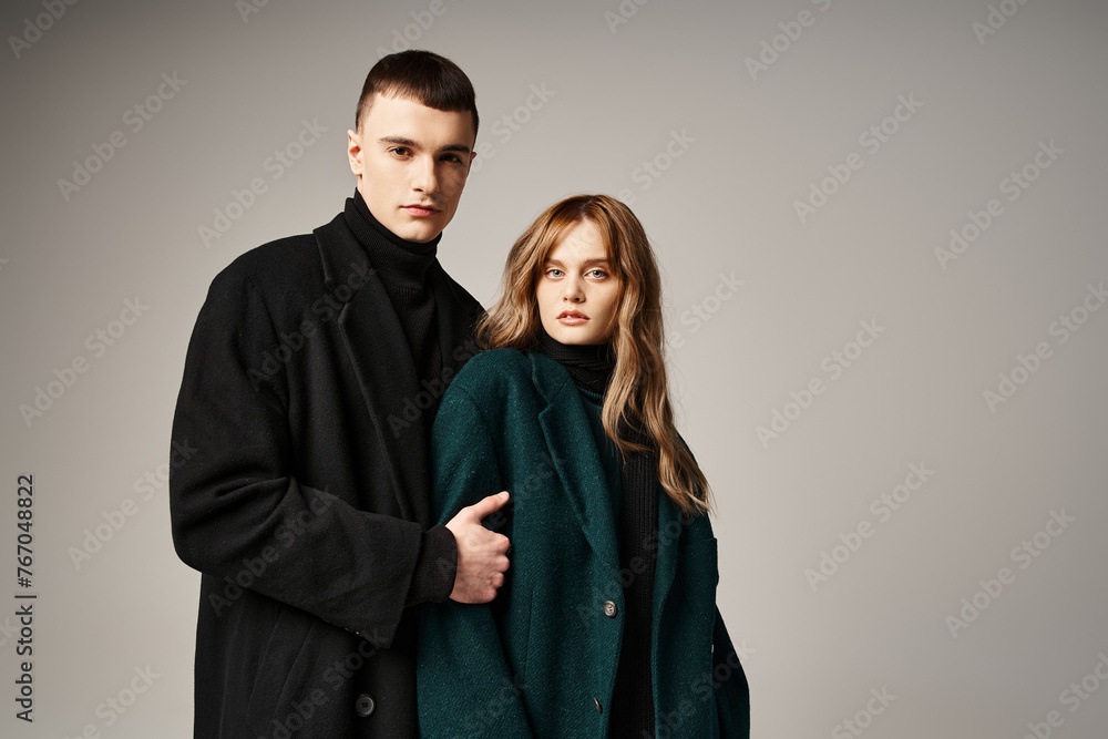 fashionable couple in stylish coats posing together on gray backdrop and looking at camera