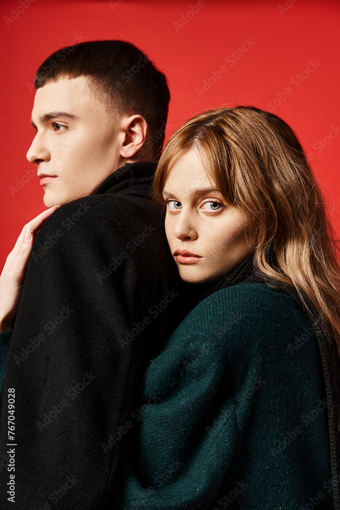 alluring woman in coat looking at camera and posing lovingly with her boyfriend on red backdrop