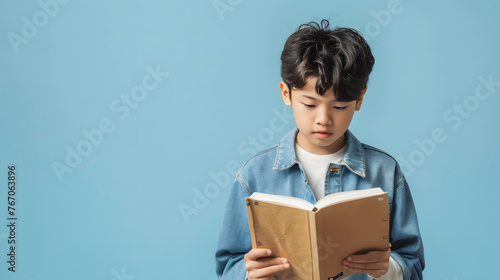 A sad Asian boy is reading a brown book on a plain blue background with copy-space for text.
