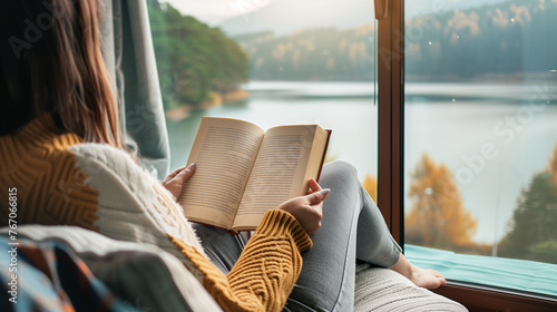 A woman is reading a book while sitting on a couch with a view of a lake. Concept of relaxation and tranquility, as the woman enjoys her book in a peaceful setting. Evening time with mountain view. photo