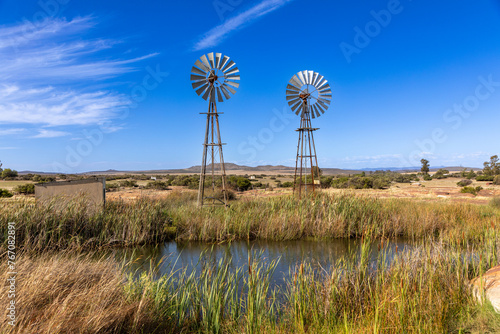Two old school metal windmills in the semi arid Karoo area of South Africa.  In the foreground is a large pool of water from the windmills.  