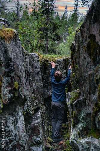 Young Explorer Scaling a Forest Crevasse in Sweden. A boy climbing carefully between towering rock walls covered in moss, surrounded by the dense greenery of a Swedish forest under summer evening sky.