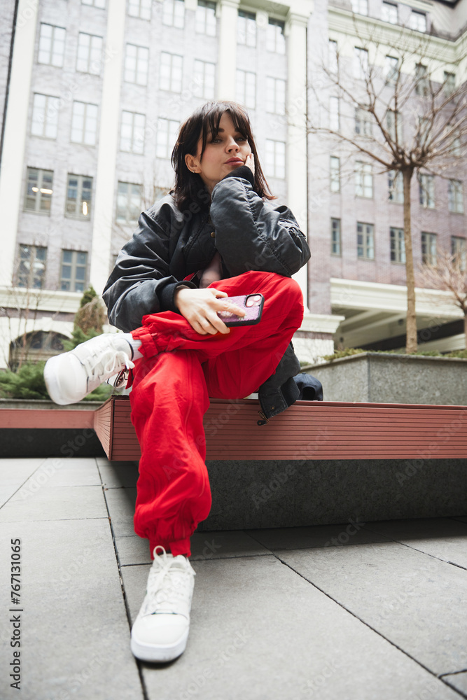 Young woman in stylish gray jacket and red pants sitting on bench with mobile phone against urban background. Concept of street style fashion, beauty, modern trends