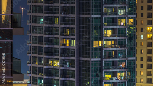 Glowing windows in multistory modern glass and metal residential building light up at night timelapse.