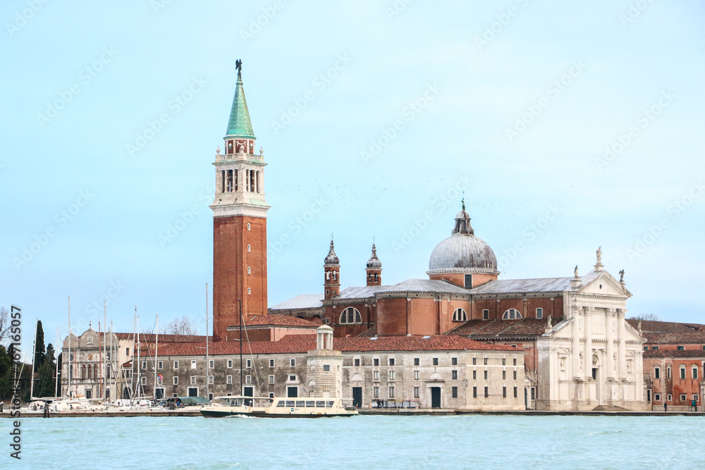 Fototapeta premium Beautiful landscape of the Church of San Giorgio Maggiore with boats docked outside, seen across the Venetian lagoon, beneath light blue sky - Venice, Italy