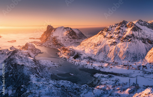 Aerial view of snowy mountains, blue sea and orange sky at sunset