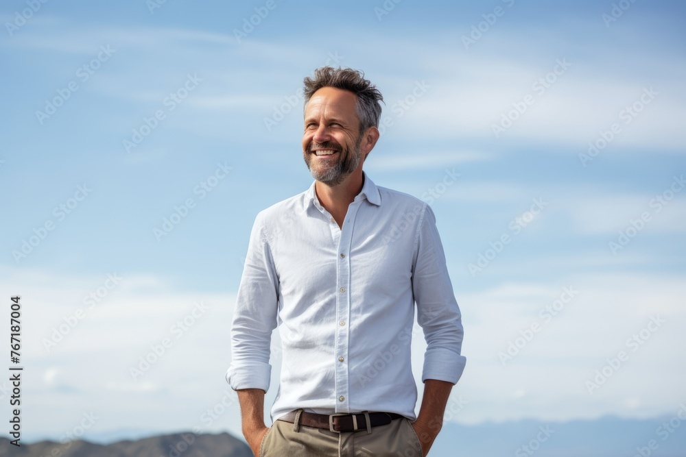 Handsome middle aged man in casual white shirt standing with hand in pocket against blue sky