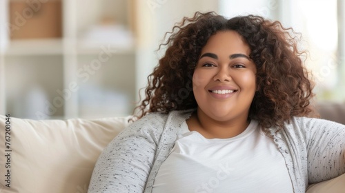Smiling woman with curly hair wearing a white top and a gray cardigan sitting on a couch with a blurred background.