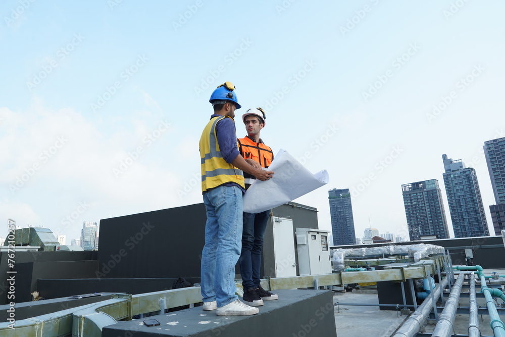 Foreman engineer and contractor engineer wearing reflective jacket, engineering helmet, holding blueprints, standing on the roof of a building under construction and behind him are tall buildings. Foreman engineer and contractor engineer wearing reflective jacket, engineering helmet, holding blueprints, standing on the roof of a building under construction and behind him are tall buildings.