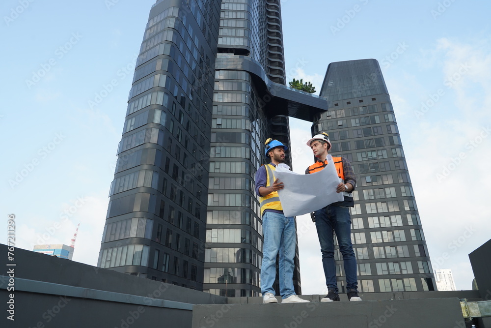 Foreman engineer and contractor engineer wearing reflective jacket, engineering helmet, holding blueprints, standing on the rooftop of a tall building in the background.