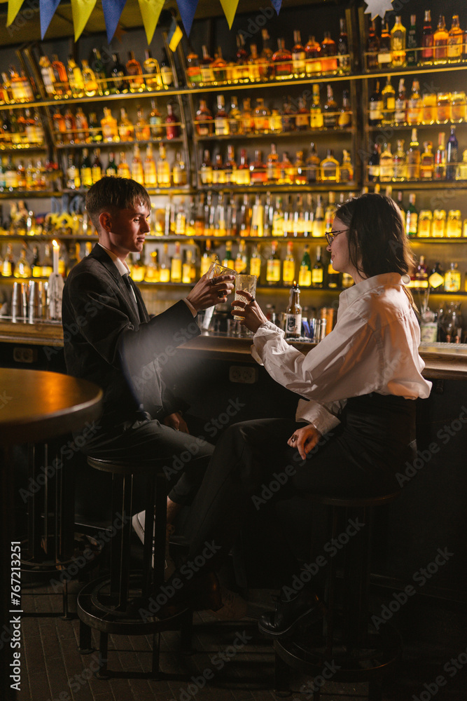 Young people greet each other and clink alcoholic drinks on holiday in modern club. Cozy evening in nightclub near patriotic bar