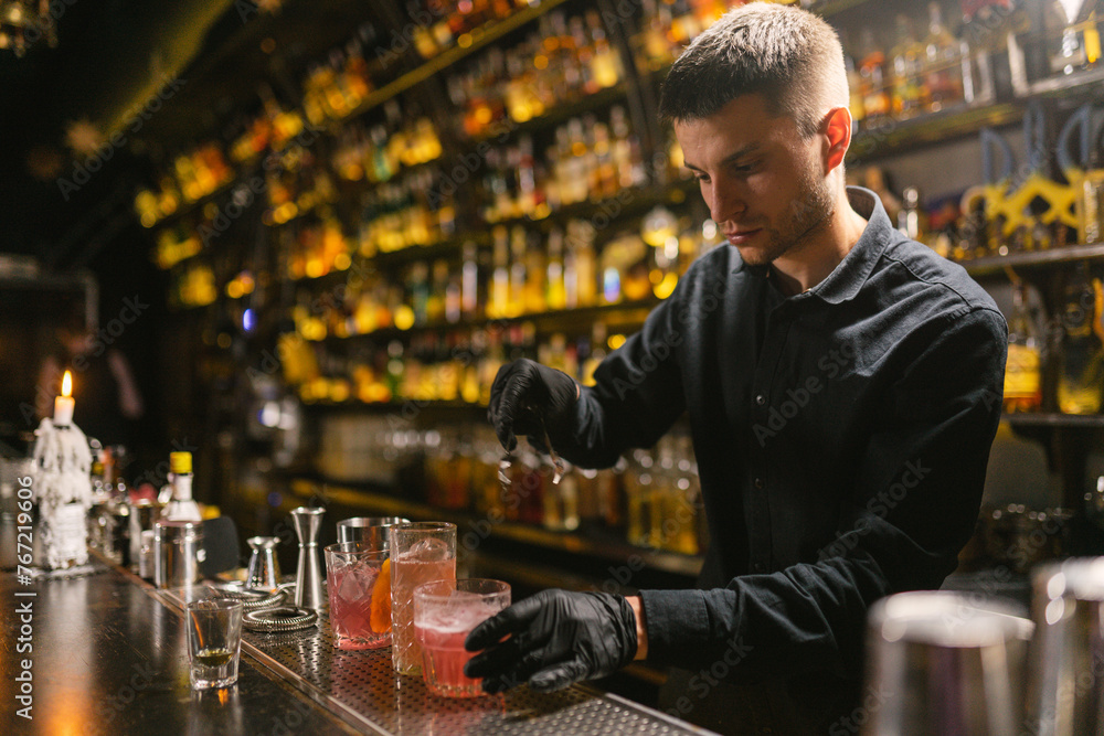 Skilled barman makes alcoholic cocktails for elegant bar guests. Barkeeper serves glasses with drinks standing on metal part of bar counter