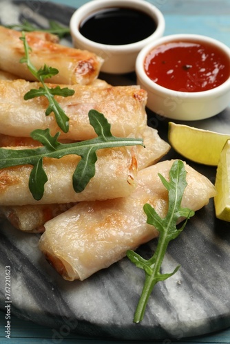 Tasty fried spring rolls, arugula, lime and sauces on light blue table, closeup