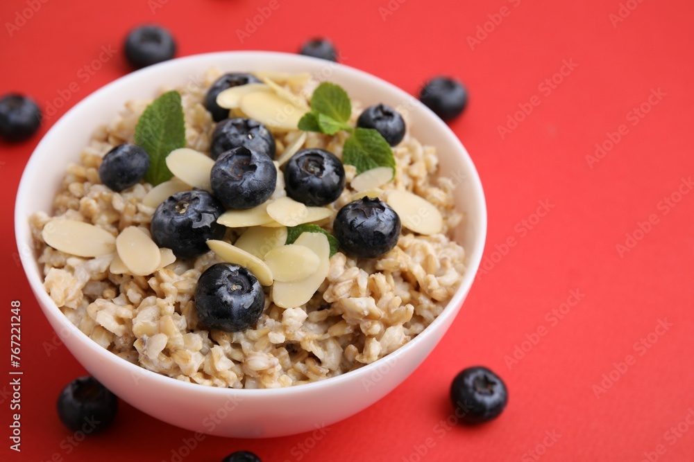 Tasty oatmeal with blueberries, mint and almond petals in bowl surrounded by fresh berries on red background, closeup. Space for text Tasty oatmeal with blueberries, mint and almond petals in bowl surrounded by fresh berries on red background, closeup. Space for text