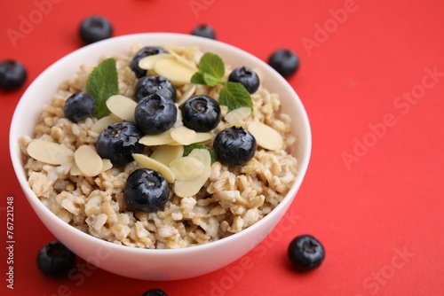 Tasty oatmeal with blueberries, mint and almond petals in bowl surrounded by fresh berries on red background, closeup. Space for text