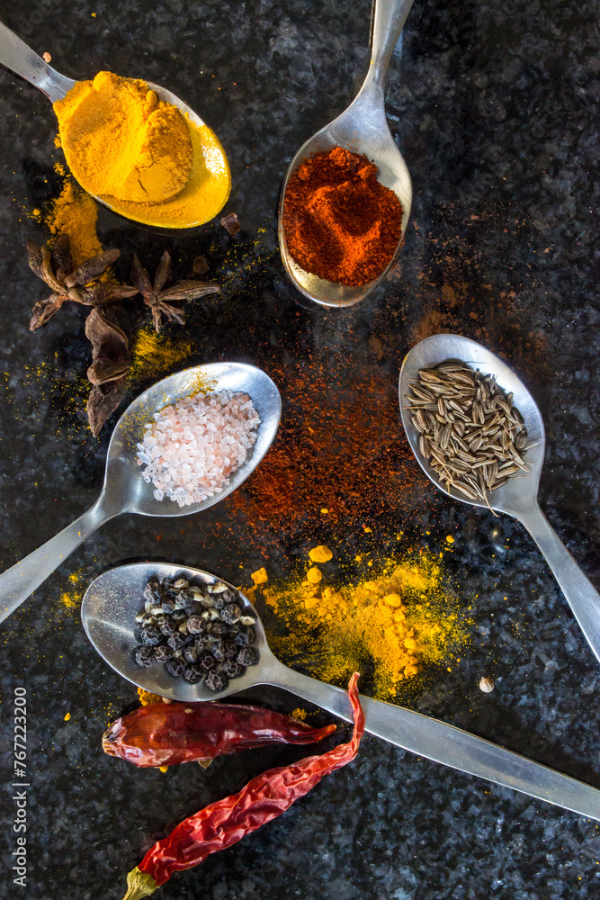 Five stainless steel teaspoons, filled with various types of spices, namely salt, pepper, cumin, turmeric and curry powder