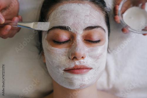 Close-up view of a beautiful young woman with closed eyes enjoying facial mask being applied at beauty spa center.