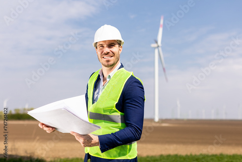 Smiling Engineer Reviewing Plans Near Wind Turbines on a Sunny Day