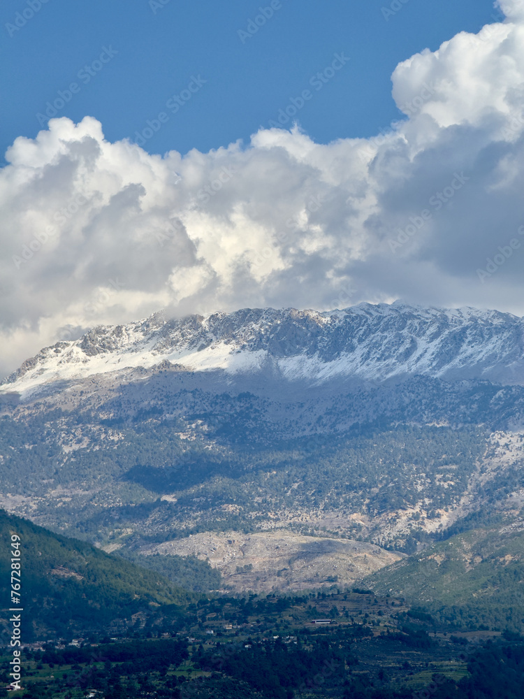 Snow-capped mountain peaks rise majestically above the lush landscape near Köprülü Canyon, framed by billowing clouds.