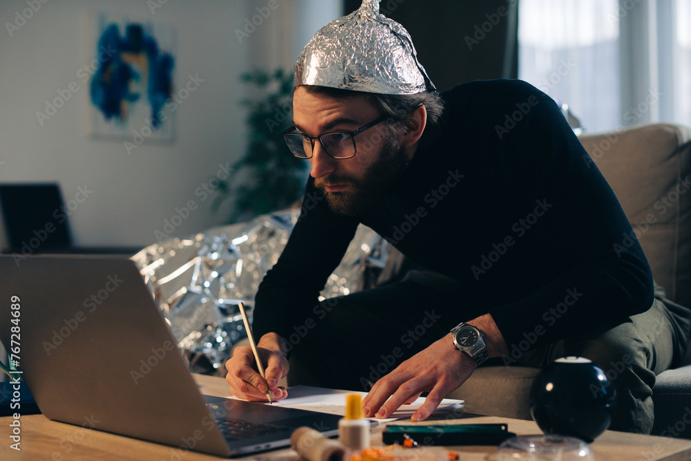 Conspiracy theory. A man in a tinfoil hat looks for signs while watching a video on a laptop. - obrazy, fototapety, plakaty 