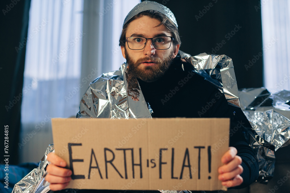 Flat Earth Advocate: Man in Tin Foil Hat and Blanket Holds Sign 'Earth is Flat - obrazy, fototapety, plakaty 