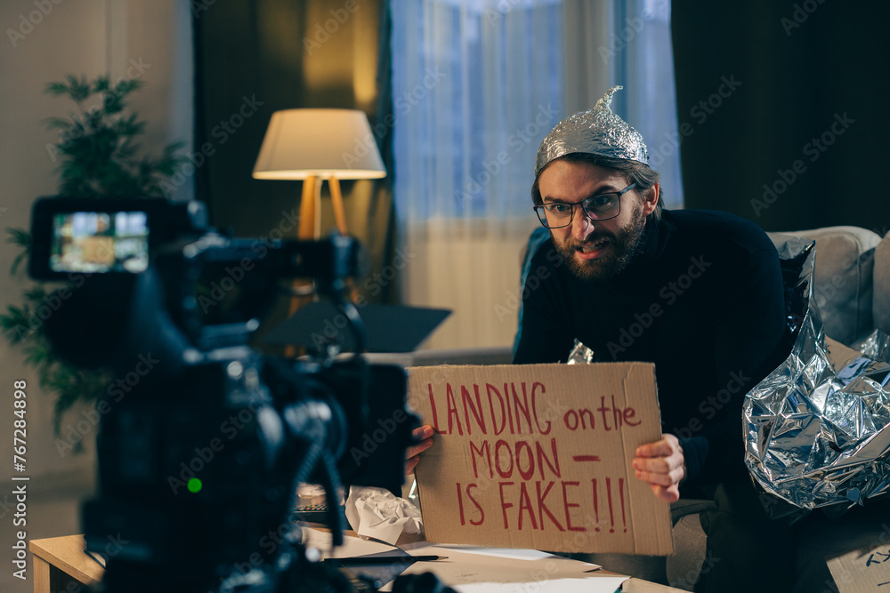 A conspiracy theorist shoots pseudoscientific videos on camera. A man in a tinfoil hat and a sign in his hands sits on a couch in front of the camera. - obrazy, fototapety, plakaty 