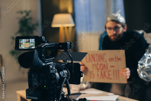 A conspiracy theorist shoots pseudoscientific videos on camera. A man in a tinfoil hat and a sign in his hands sits on a couch in front of the camera. photo